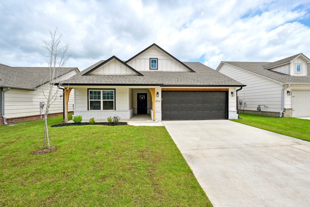 A modern one-story house with a two-car garage and a long paved driveway, an overhanging porch, and a lush lawn with a you...