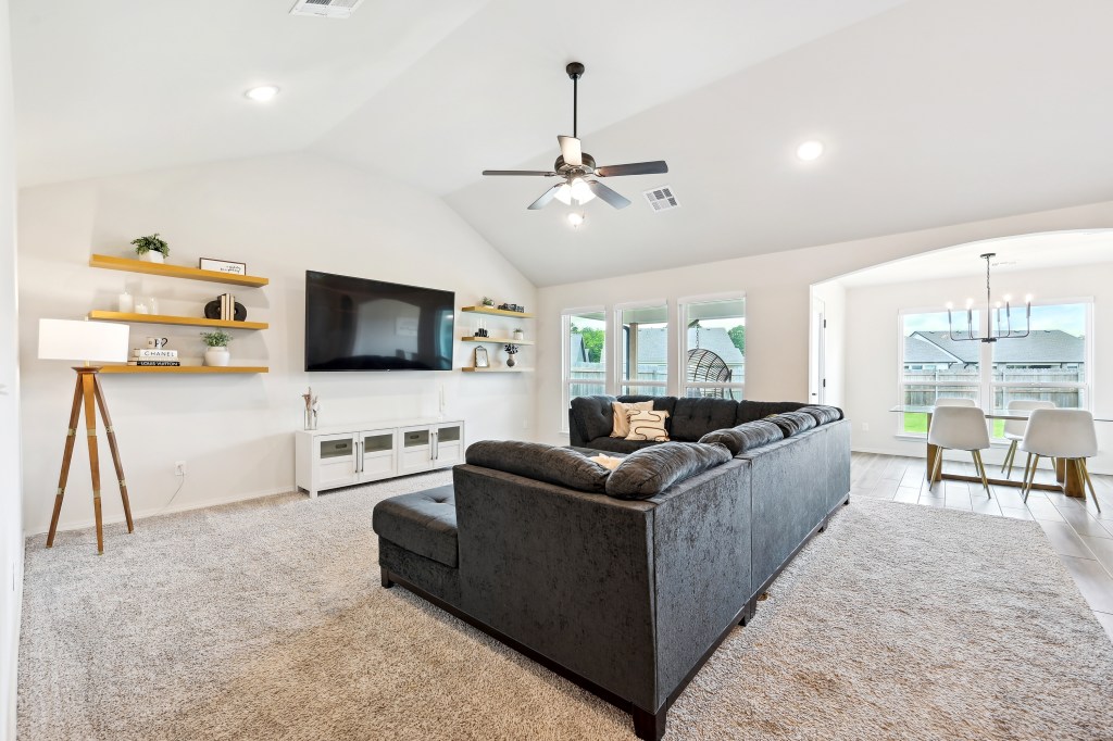 A modern living room with a gray couch and neutral decor, featuring a large TV mounted on the wall, a dining area with a t...