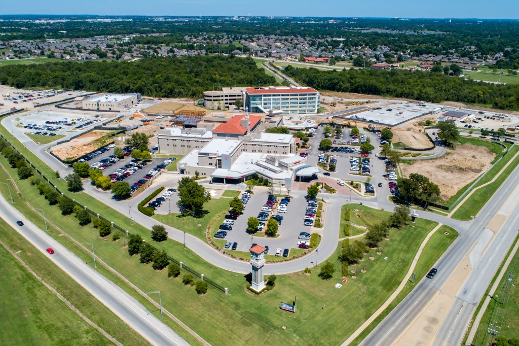 An aerial view of a large white building with a red roof, surrounded by a parking lot, tree-lined streets, and a highway w...