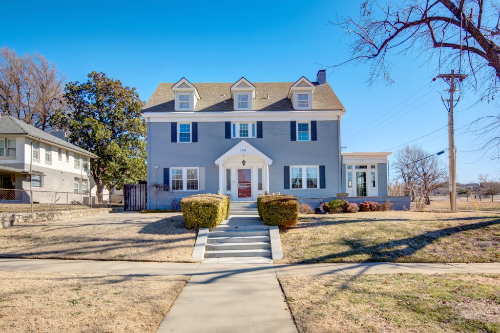 A large two-story house with a light gray exterior and white trim, featuring multiple windows and a covered front porch wi...